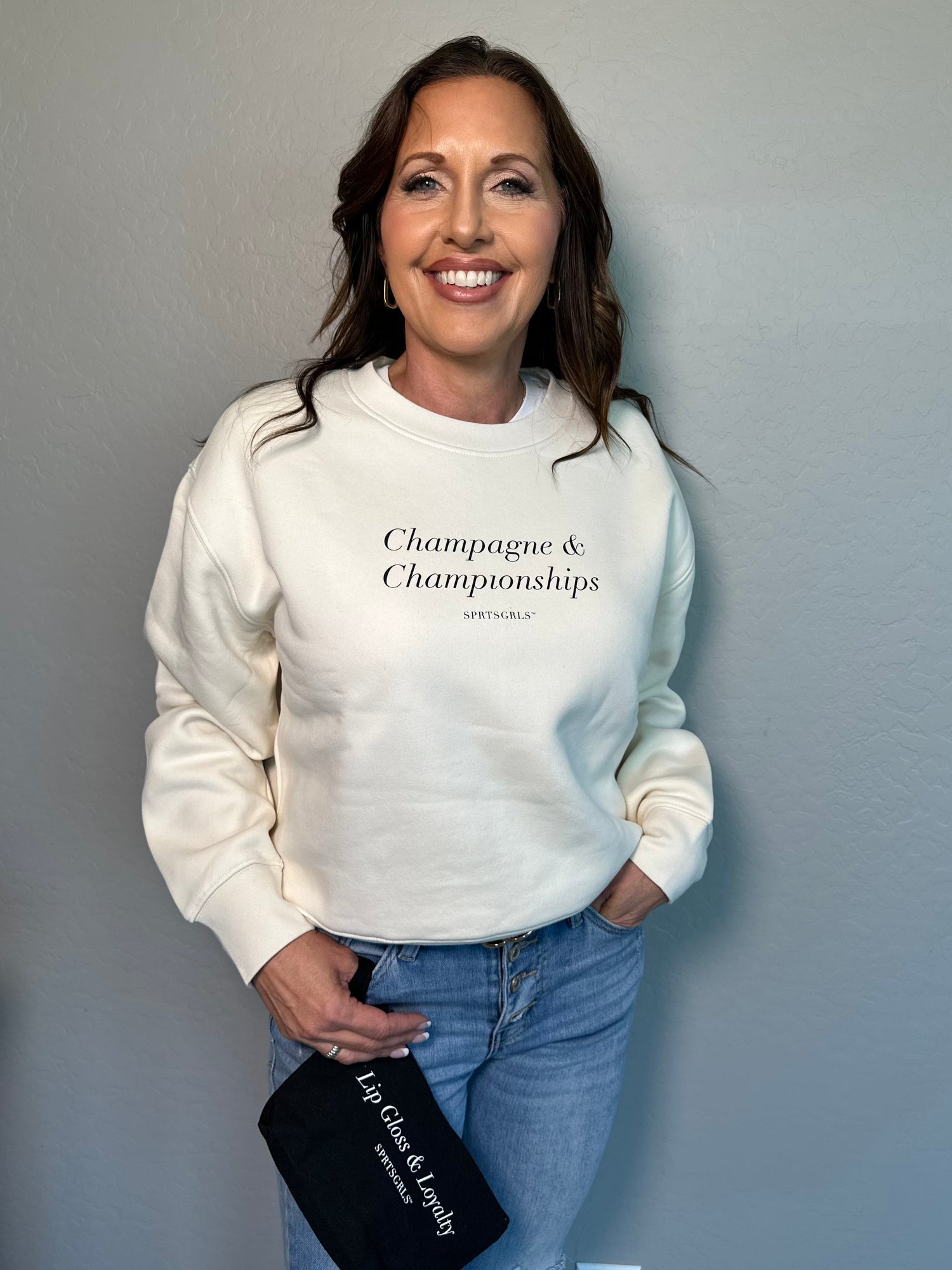 SPRTSGRLS owner Amy Cannatta is wearing a cream sweatshirt with the text 'Champagne & Championships' printed on the chest, paired with light blue jeans, and holding a black clutch with the slogan 'Lip Gloss & Loyalty'.