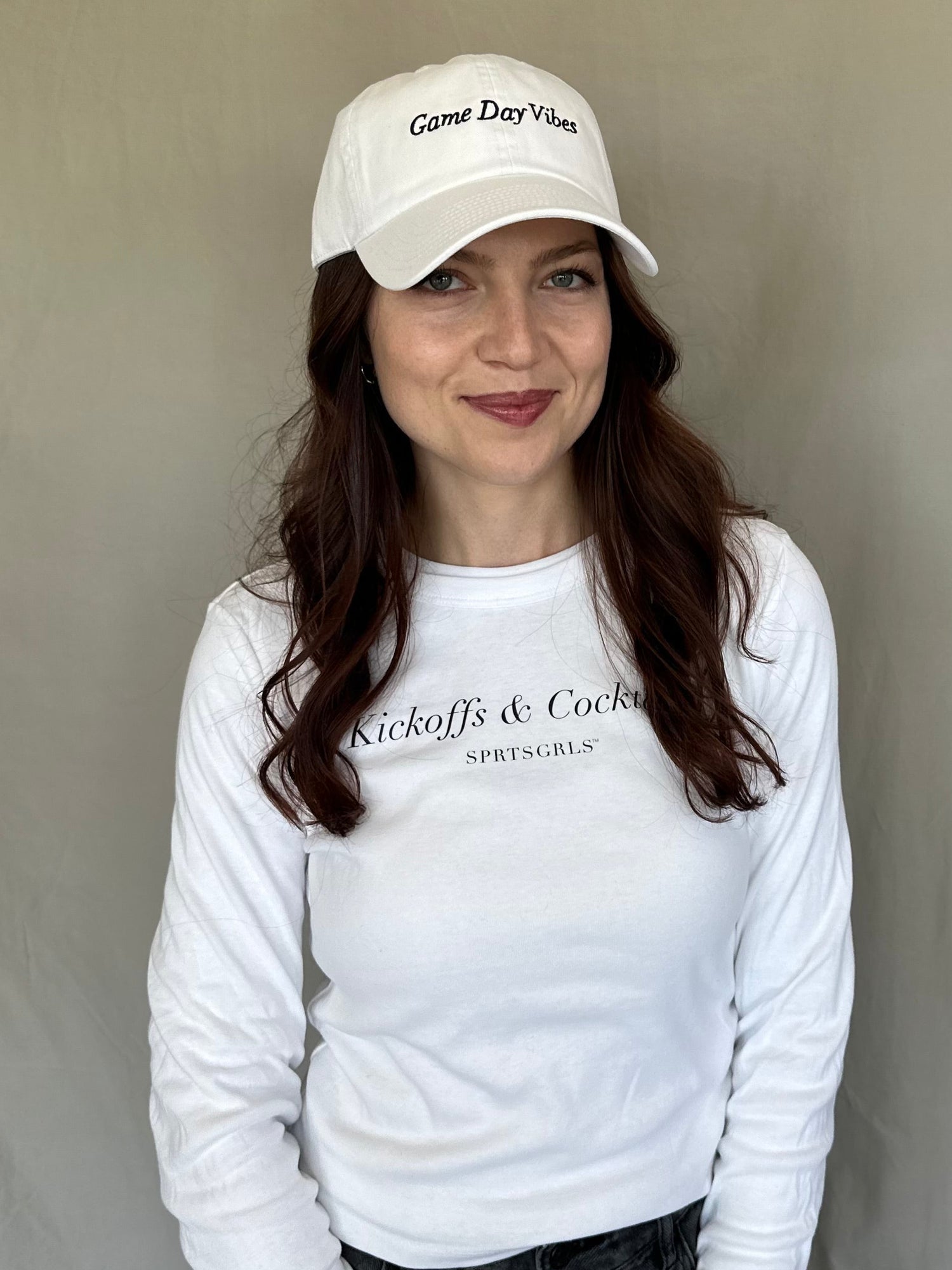 A woman wearing a white long-sleeve crewneck shirt with 'Kickoffs & Cocktails' printed on the chest, paired with a white baseball cap that reads 'Game Day Vibes'.