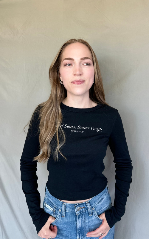 A woman with long light brown hair wearing a black long-sleeve crewneck t-shirt with white text that reads 'Seats Better Outfit' and 'SAPPHIRE', paired with blue jeans, standing against a neutral beige backdrop.