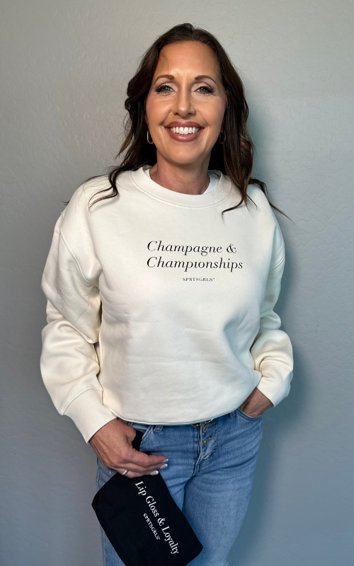 A woman smiling while wearing a cream-colored crewneck sweatshirt with the text 'Champagne & Championships' printed on the chest. She is also wearing blue jeans and holding a black clutch bag with white text that reads 'Lip gloss & Loyalty'.